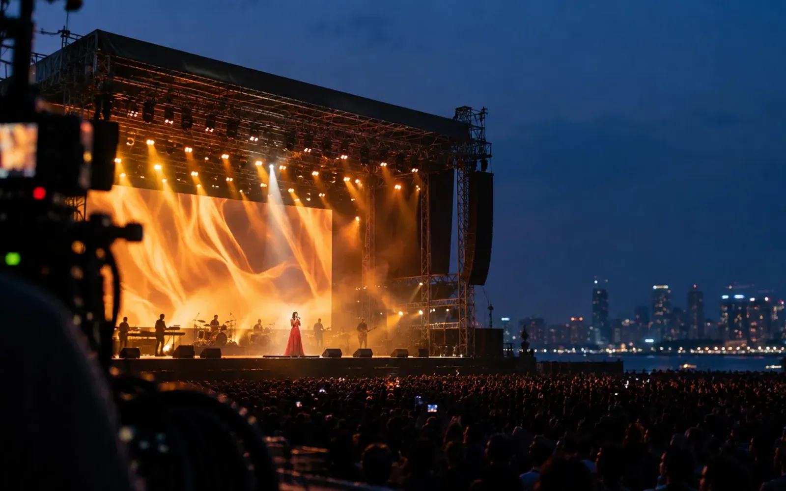 An open-air concert at night: a singer in red before a flame-wall LED screen, the crowd and a lit skyline across the water