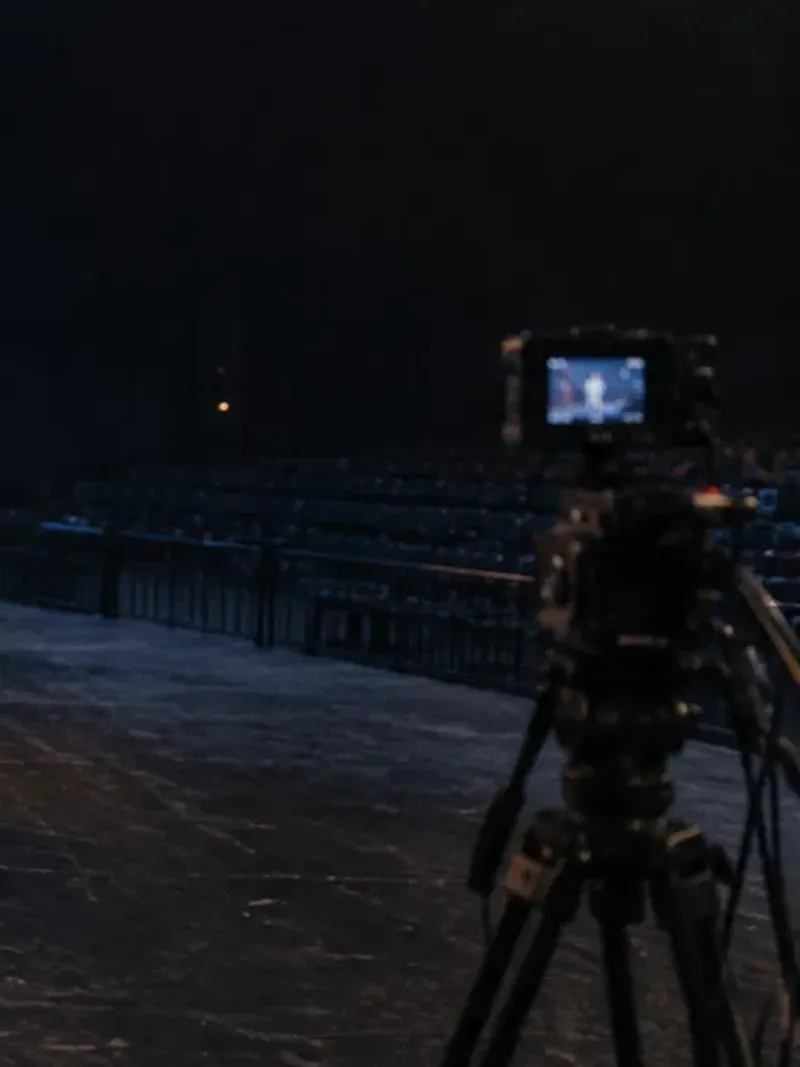 A camera on a tripod, its monitor glowing, facing rows of empty seats in a dark arena
